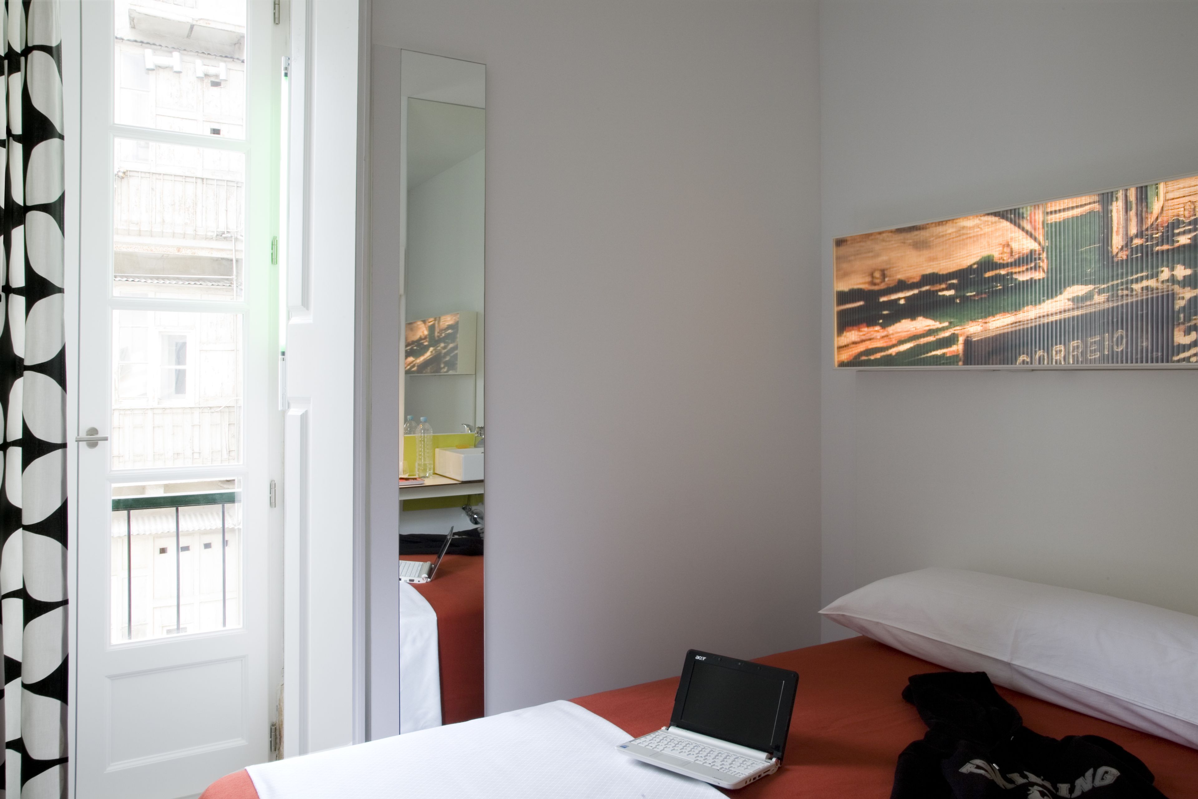 A bedroom with a bed, a laptop on the bed, a pillow, a wall-mounted painting, and a door with a view of the outside.