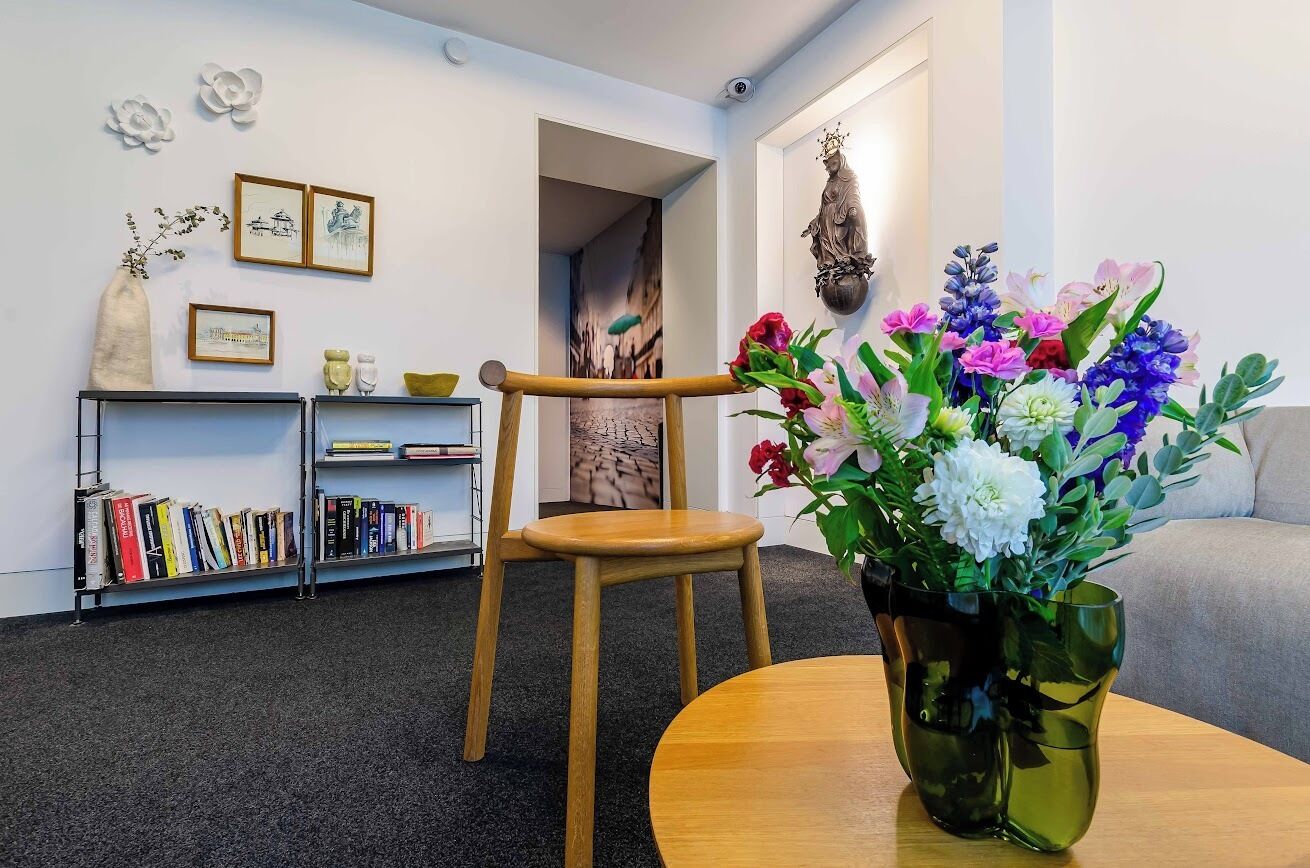 Ein Zimmer mit einem Holzsessel, einem runden Tisch mit Blumen in einer Vase, einem Bücherregal mit Büchern und einer Wand mit gerahmten Bildern und einer Statue.