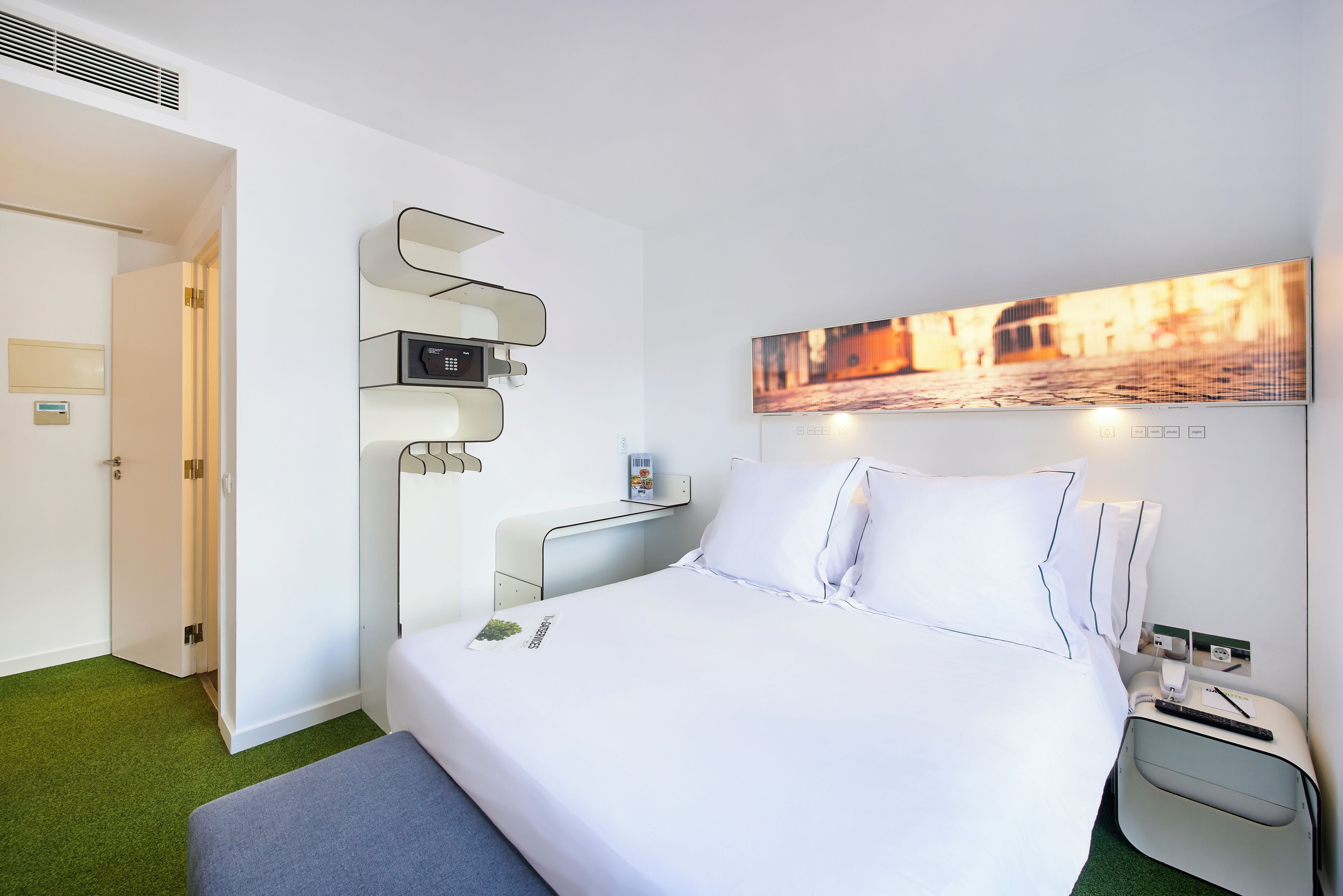 A hotel room with a bed, bedside table, and a wall-mounted control panel.