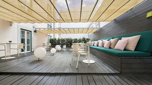Un área de estar al aire libre con una terraza de madera, un sofá verde con cojines rosas y una fila de sillas blancas.