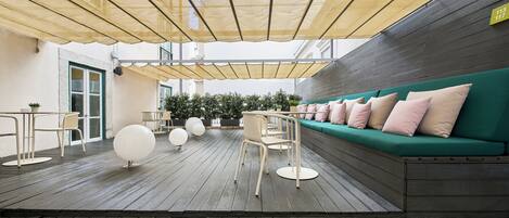 An outdoor seating area with a wooden deck, a green sofa with pink cushions, and a row of white chairs.