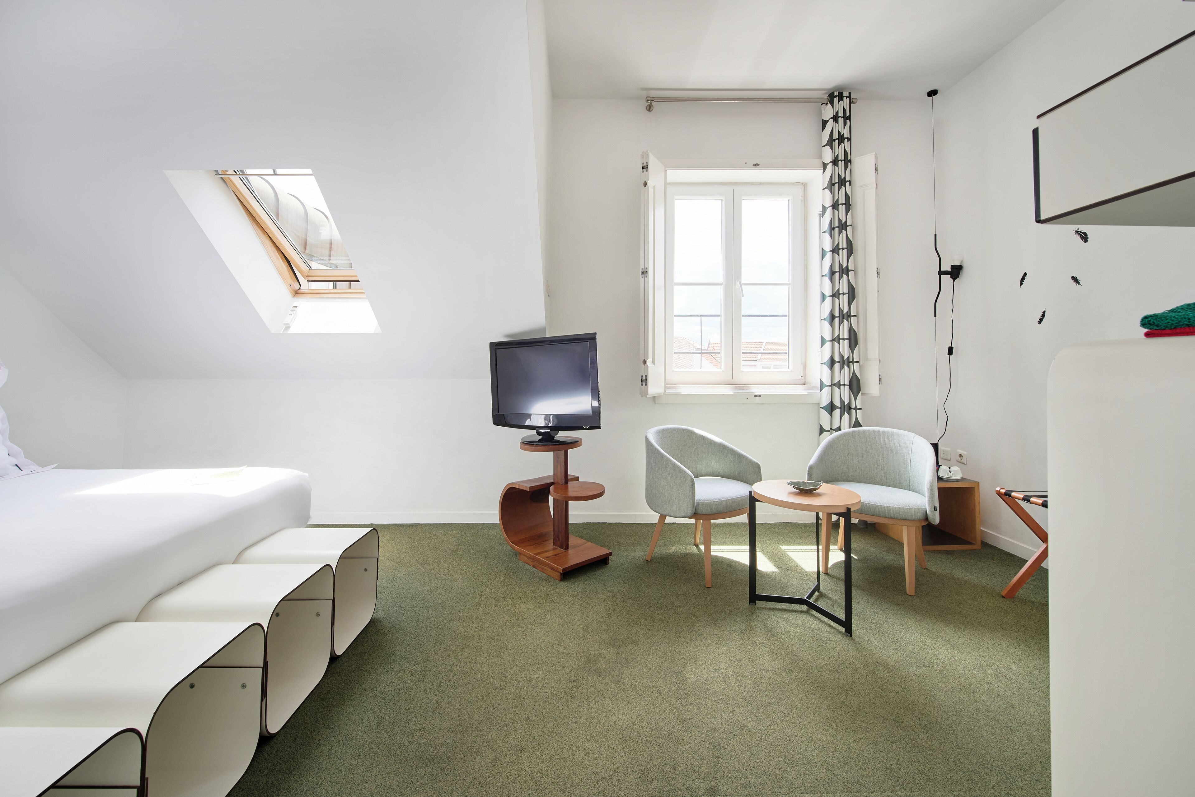 Habitación de hotel moderna con una cama, televisor, dos sillones, una mesita, ventana con cortinas e iluminación en el techo.