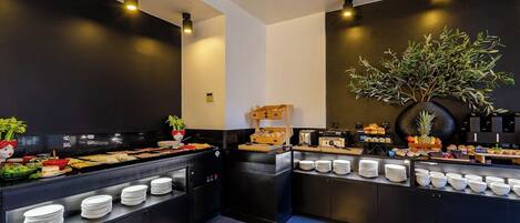 Montaje tipo bufé con diversos alimentos y platos, una planta grande en maceta y una cocina moderna.