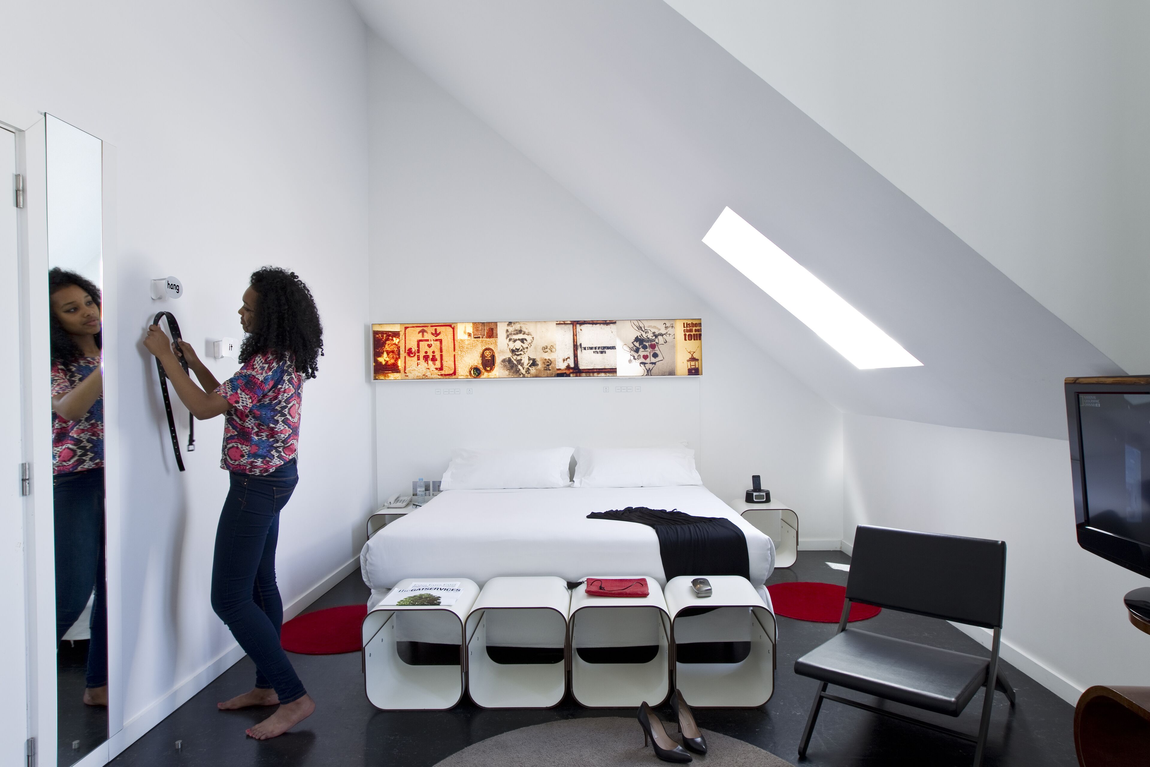 A modern hotel room with a bed, a TV, a mirror, and a series of white storage units.