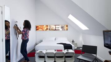 A modern hotel room with a bed, a TV, a mirror, and a series of white storage units.