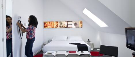 A modern hotel room with a bed, a TV, a mirror, and a series of white storage units.