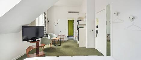A modern hotel room with a flat-screen TV, a bed, a desk, and a chair. There is a green carpet and a white wall with a door and a mirror.