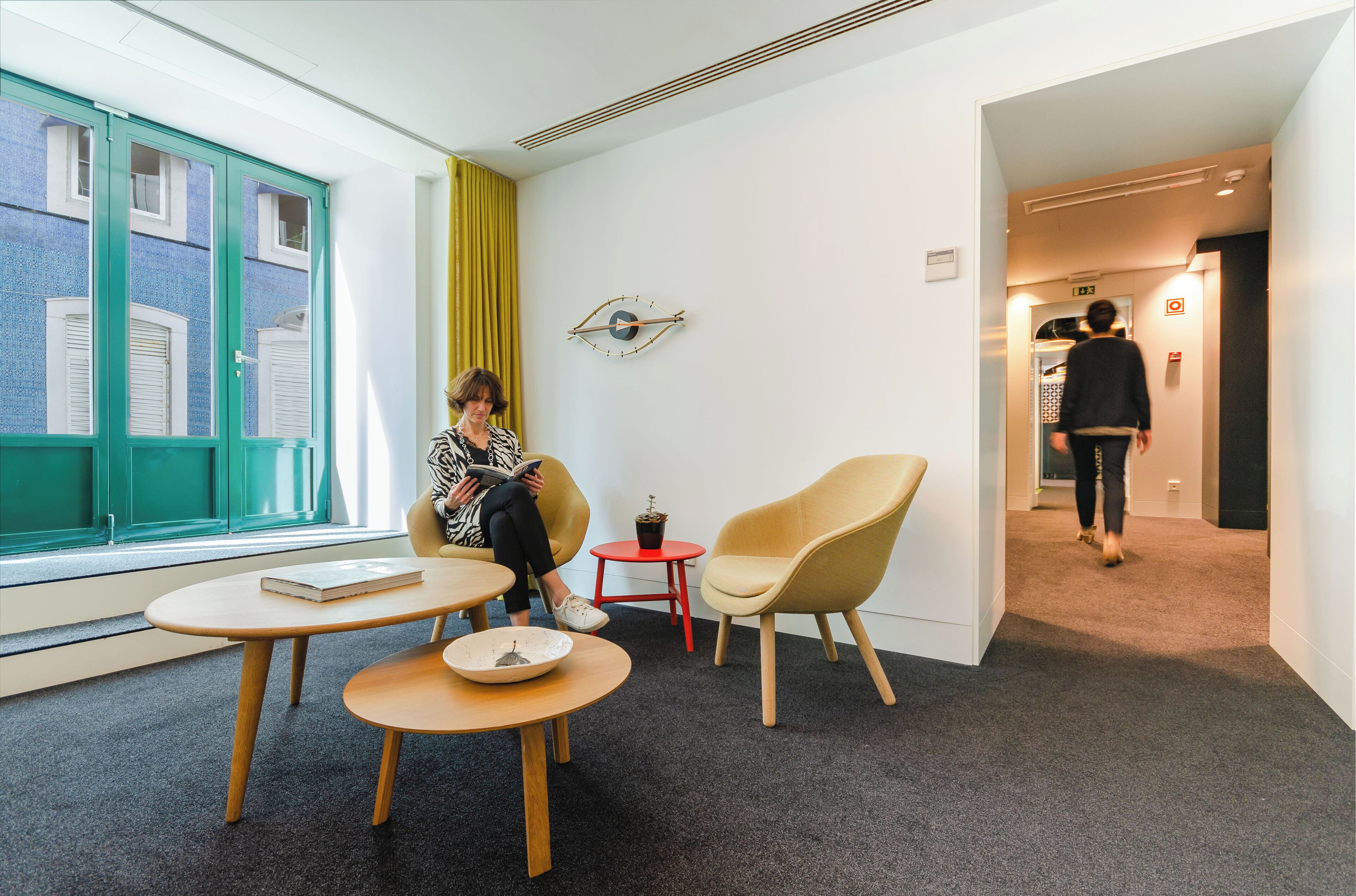 Un hall d’hôtel moderne avec une grande fenêtre, un fauteuil jaune, une petite table avec un livre et une horloge murale originale.