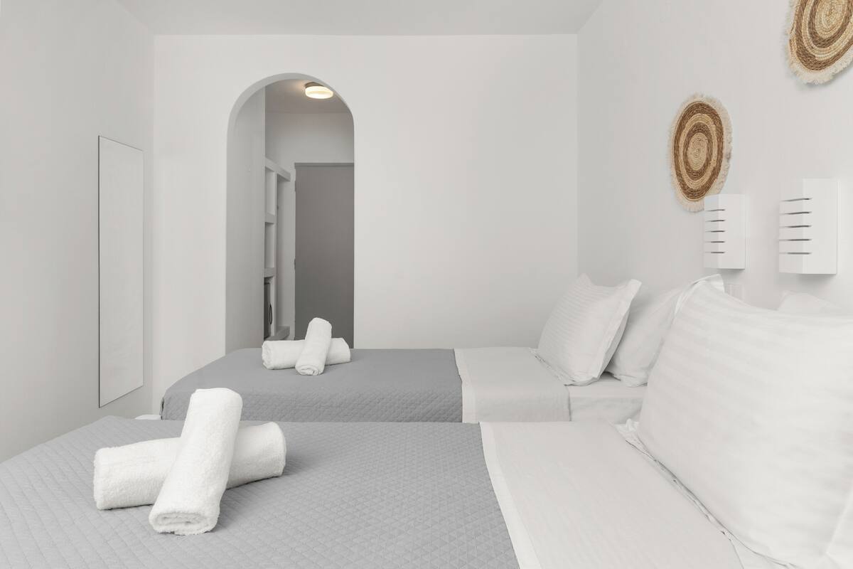 A modern hotel room with two beds, a large mirror, a small bathroom, and a view through an arched doorway.