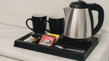 Una tetera de acero inoxidable, dos tazas negras, una cuchara y una bandeja con bolsitas de té sobre una encimera de mármol.