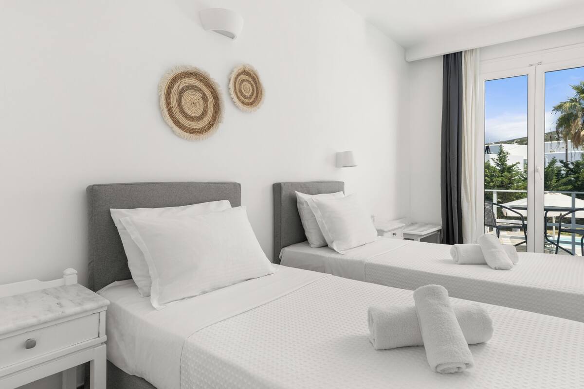 A hotel room with two beds, a nightstand, a lamp, and a view of the outdoors.