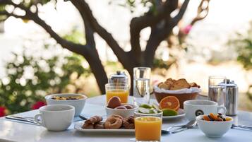 Una mesa de desayuno con diversas comidas y bebidas, ubicada al aire libre con árboles de fondo.