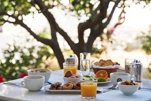 A breakfast table with various food items and drinks, set outdoors with trees in the background.