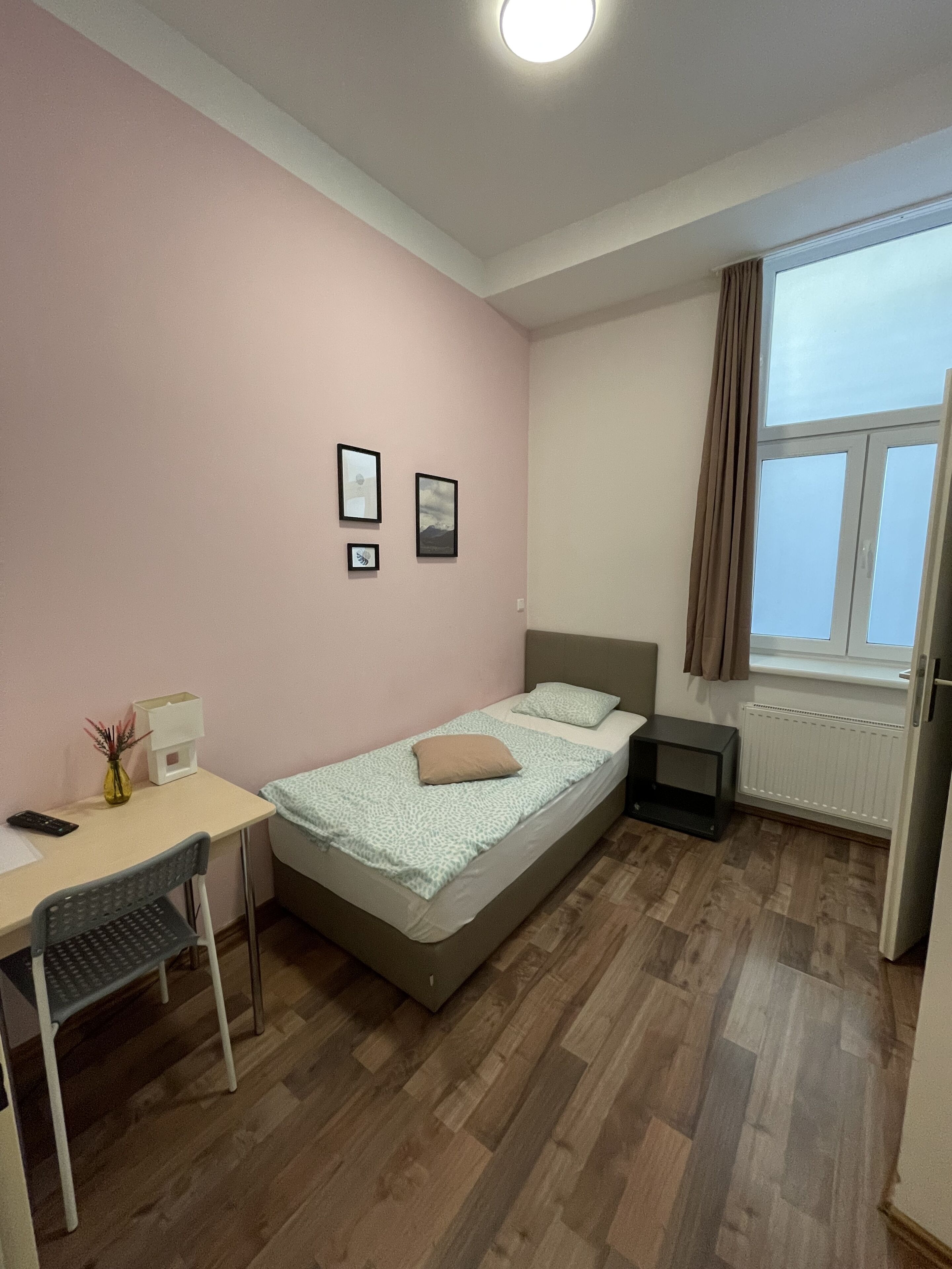 Un dormitorio pequeño y ordenado con una cama individual, un escritorio y una silla. Hay dos cuadros en la pared, un radiador y una ventana con cortinas.
