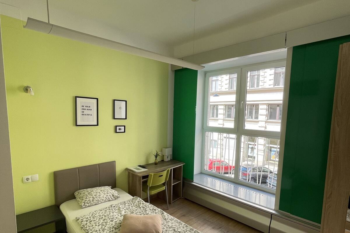 Ein Schlafzimmer mit einem Bett, einem Schreibtisch und einem Fenster mit Blick auf Gebäude und Autos.