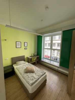 A bedroom with a bed, a desk, and a window with a view of buildings and cars.