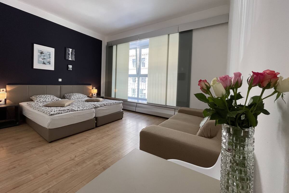 Ein modernes Schlafzimmer mit Bett, einer Couch, einer Vase mit Blumen und einem Fenster mit Jalousien.