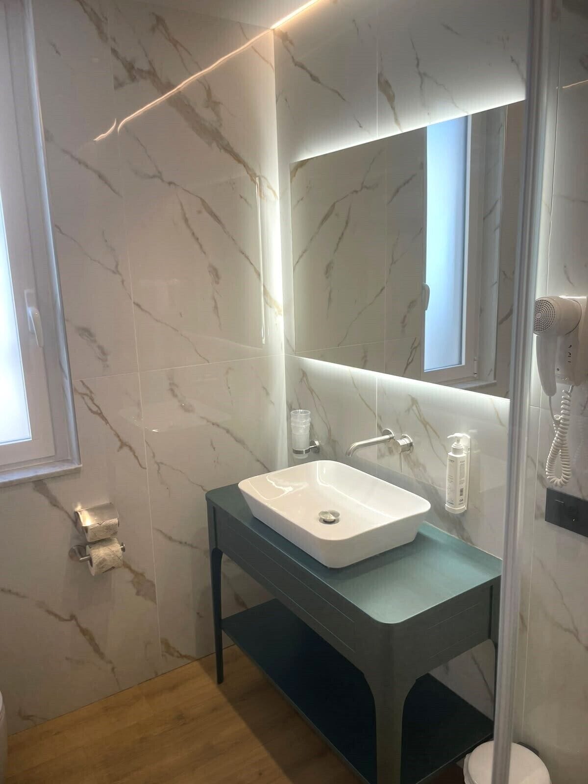 Une salle de bain avec un mur en marbre, un lavabo blanc, un miroir, un distributeur de serviettes et une douchette à main.