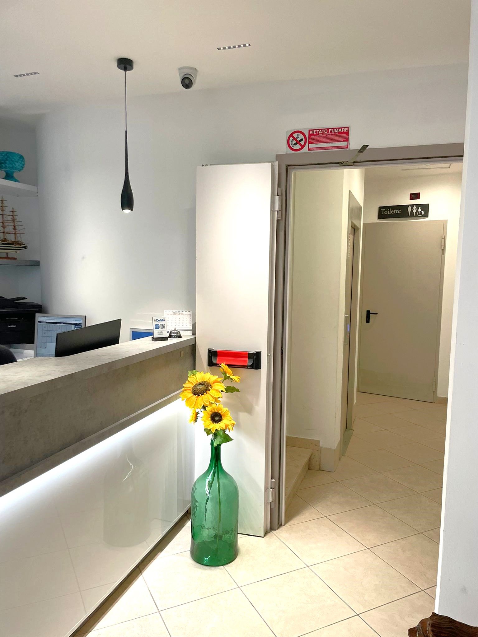 A reception area with a desk, a vase with sunflowers, a sign indicating a toilet, and a door leading to a hallway.