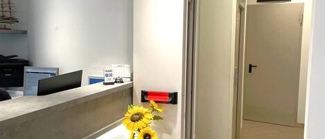 A reception area with a desk, a vase with sunflowers, a sign indicating a toilet, and a door leading to a hallway.