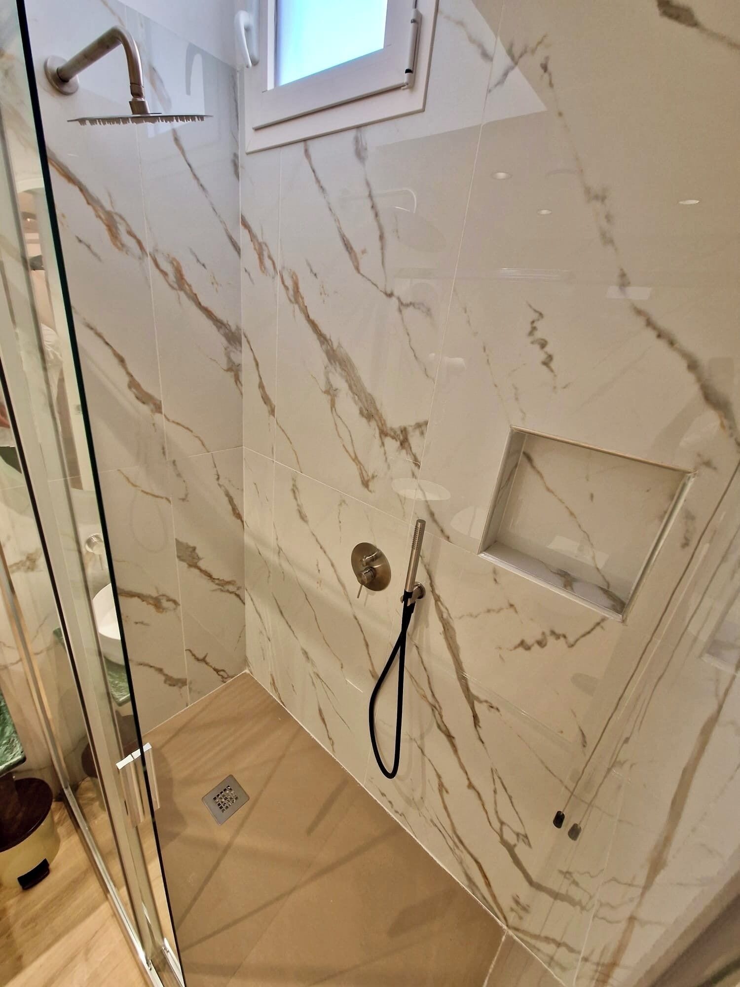 A glass-enclosed shower with marble walls, a small window, and a built-in shelf.