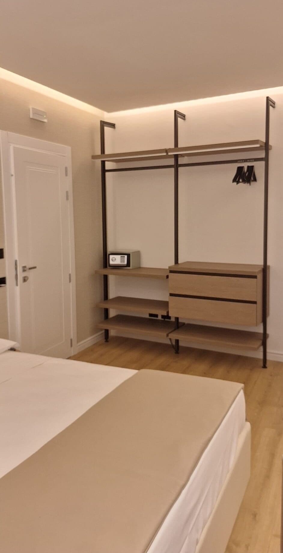 Ett sovrum med en säng, en garderob med hyllor, ett skrivbord och en liten elektronisk apparat på hyllan.