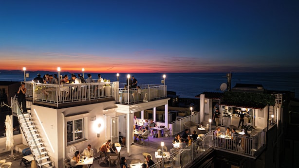 Un bar en la azotea con mesas al aire libre, vistas al mar, varias zonas de comedor y una escalera que lleva al nivel superior.