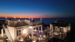 A rooftop bar with outdoor seating overlooking the sea, multiple dining areas, and a staircase leading to the upper level. - Hotel La Giara (Cefalù)