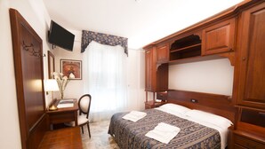 Una habitación de hotel con una cama, un escritorio con silla, un televisor montado en la pared y un armario de madera.