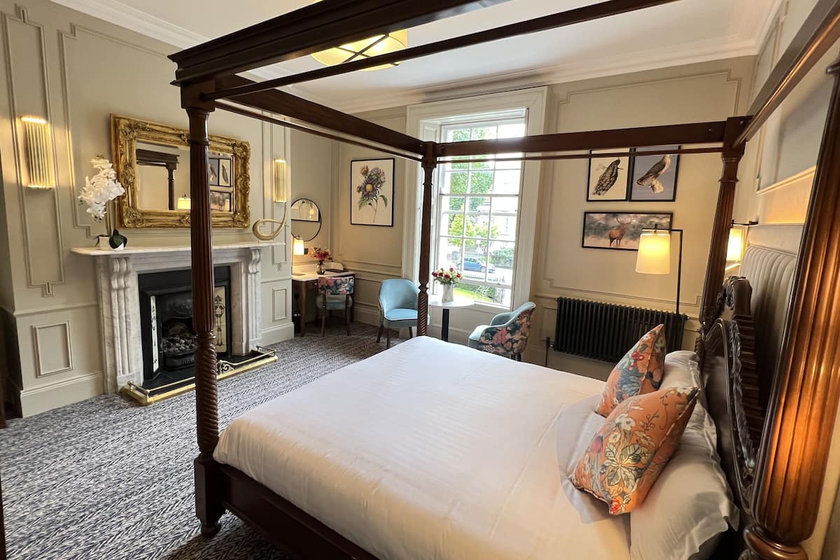 A four-poster bed with a floral pillow, a fireplace, a desk with a chair, and artwork on the walls.