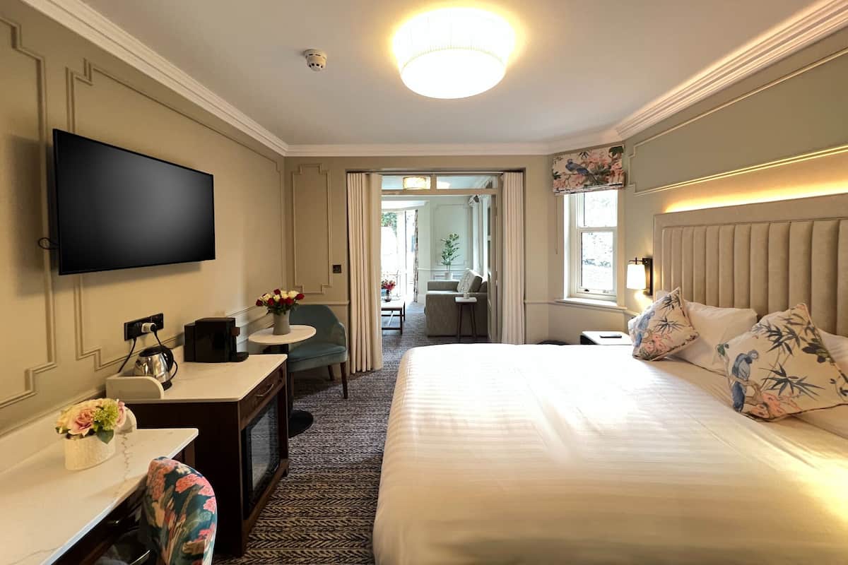 A hotel room with a flat-screen TV, a desk with a vase of flowers, a bed with floral pillows, and a view of the dining area.