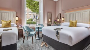A hotel room with two beds, a sitting area with a chair and table, a window with a view of buildings, and a mirror on the wall. - Waterloo Townhouse & Suites (Dublin)
