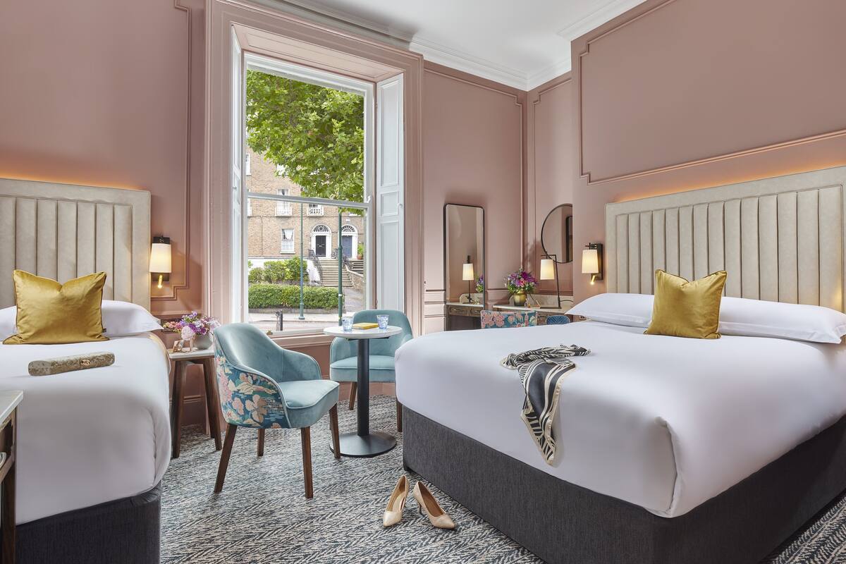 A hotel room with two beds, a sitting area with a chair and table, a window with a view of buildings, and a mirror on the wall.