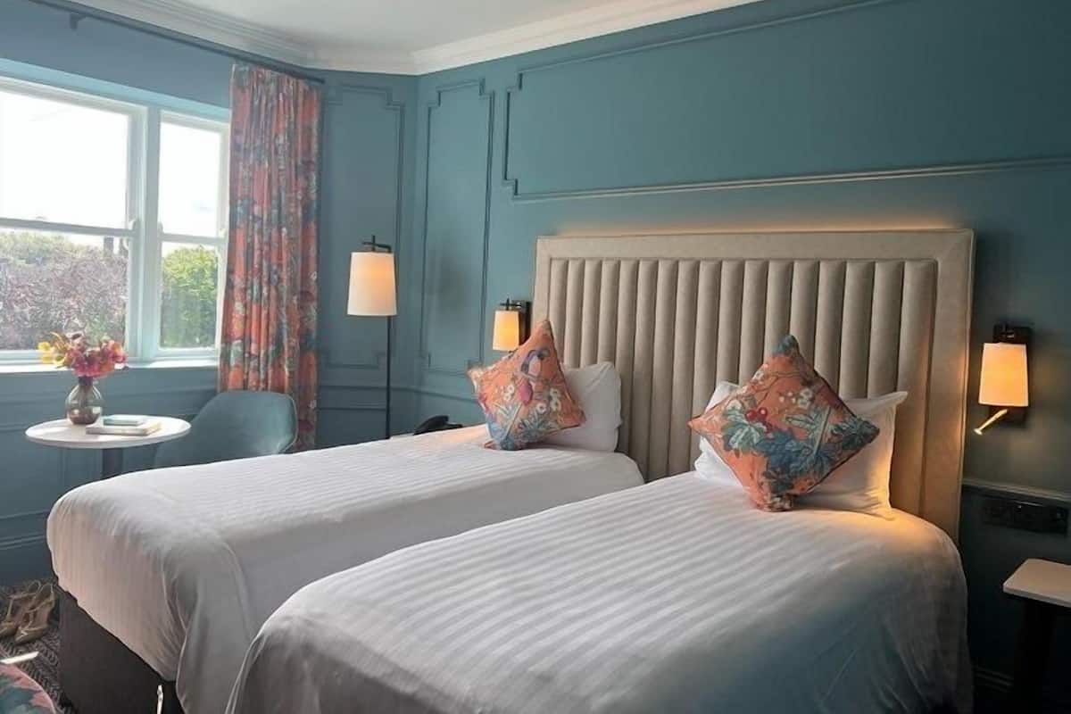 A hotel room with two beds, a window with curtains, a small table with a vase, and a lamp on the nightstand.