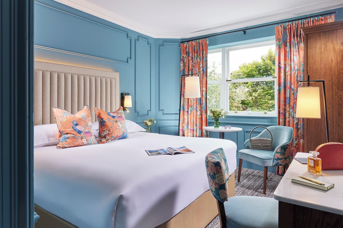 A hotel room with a bed, a desk, a chair, a lamp, and a window with curtains.