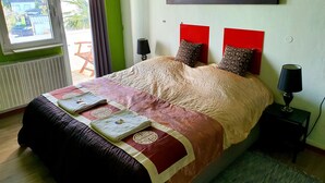 A neatly made bed with a patterned bedspread and pillows, a window with curtains, a wall-mounted painting of a mountain, and a small bedside table with a lamp. - StayInCologne (Cologne)