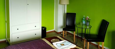 A room with a green wall, a white wardrobe, a purple bedspread, a table with chairs, and a ceiling light fixture.