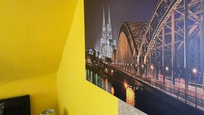 A room with a yellow wall, a framed picture of a bridge, a small table with a lamp, and a patterned wall. - StayInCologne (Cologne)