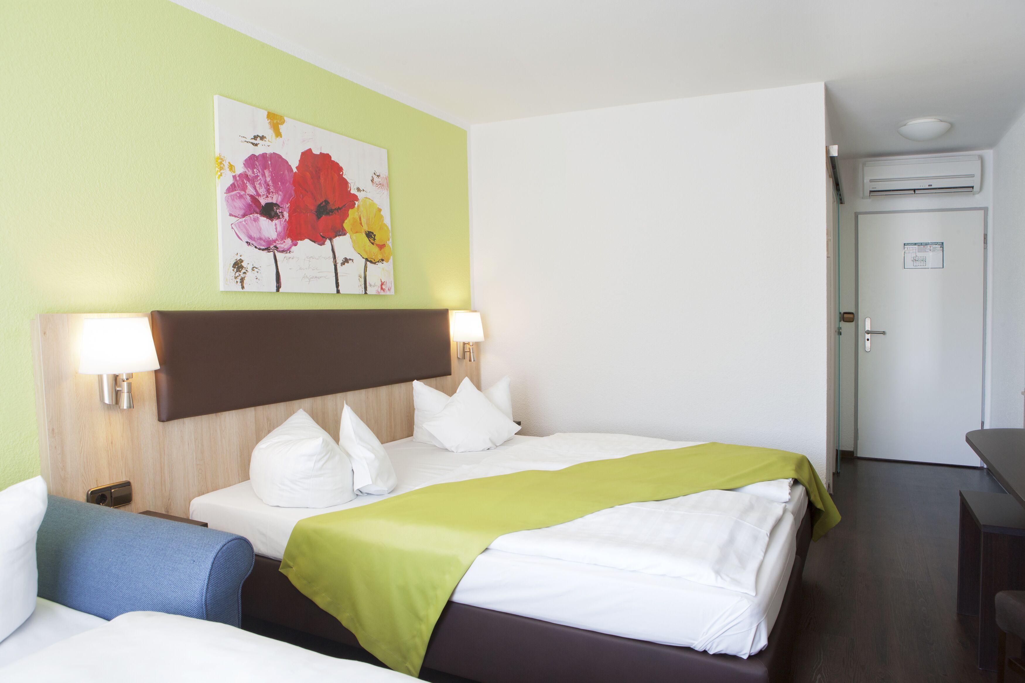 A hotel room with a bed, a desk, and a chair. There is a painting on the wall and a door to the right.