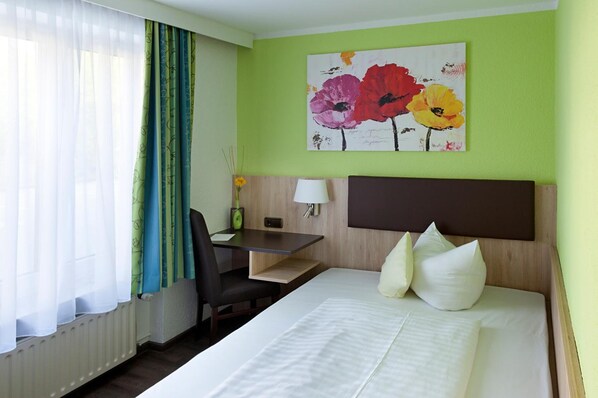 A hotel room with a bed, a desk, a chair, a lamp, and a painting of flowers on the wall.