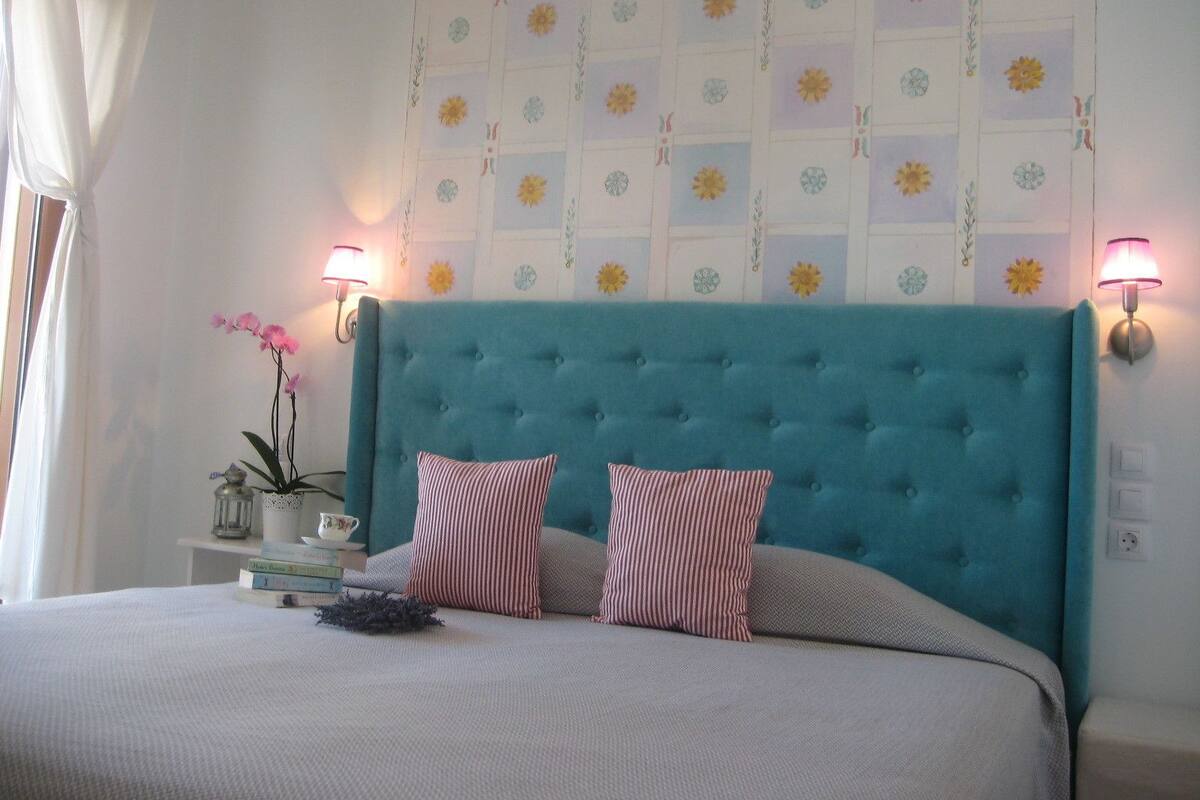 A bedroom with a teal upholstered headboard, two striped pillows, a bedside table with a lamp, and a floral-patterned wall.