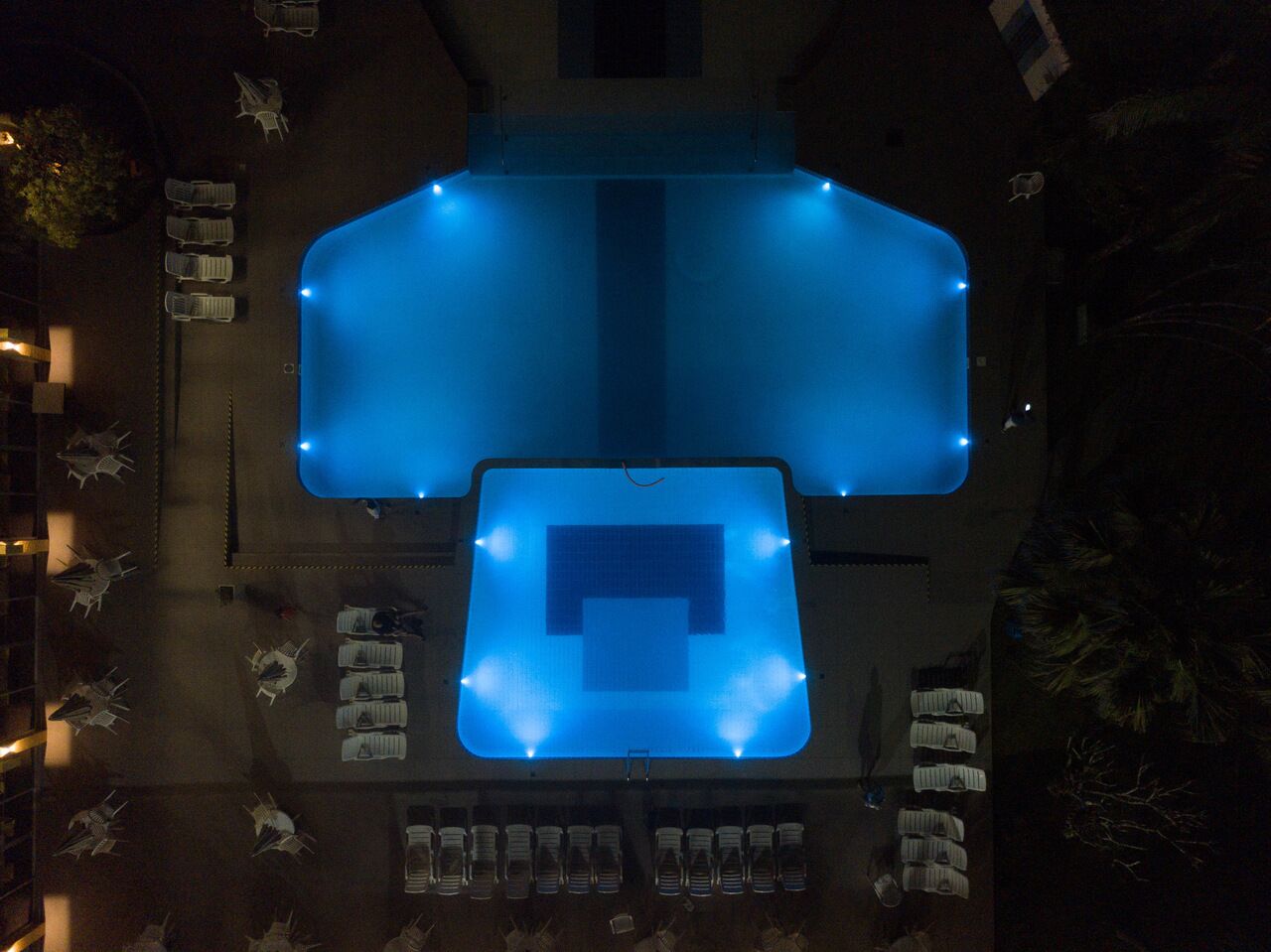 Ein Luftbild eines Hotels mit großem Schwimmbecken, angeordneten Sitzgelegenheiten und einem Parkplatz.