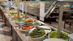 Una barra de buffet con diversas opciones de comida, incluyendo ensaladas y verduras, en un comedor bien iluminado con mesas y sillas.