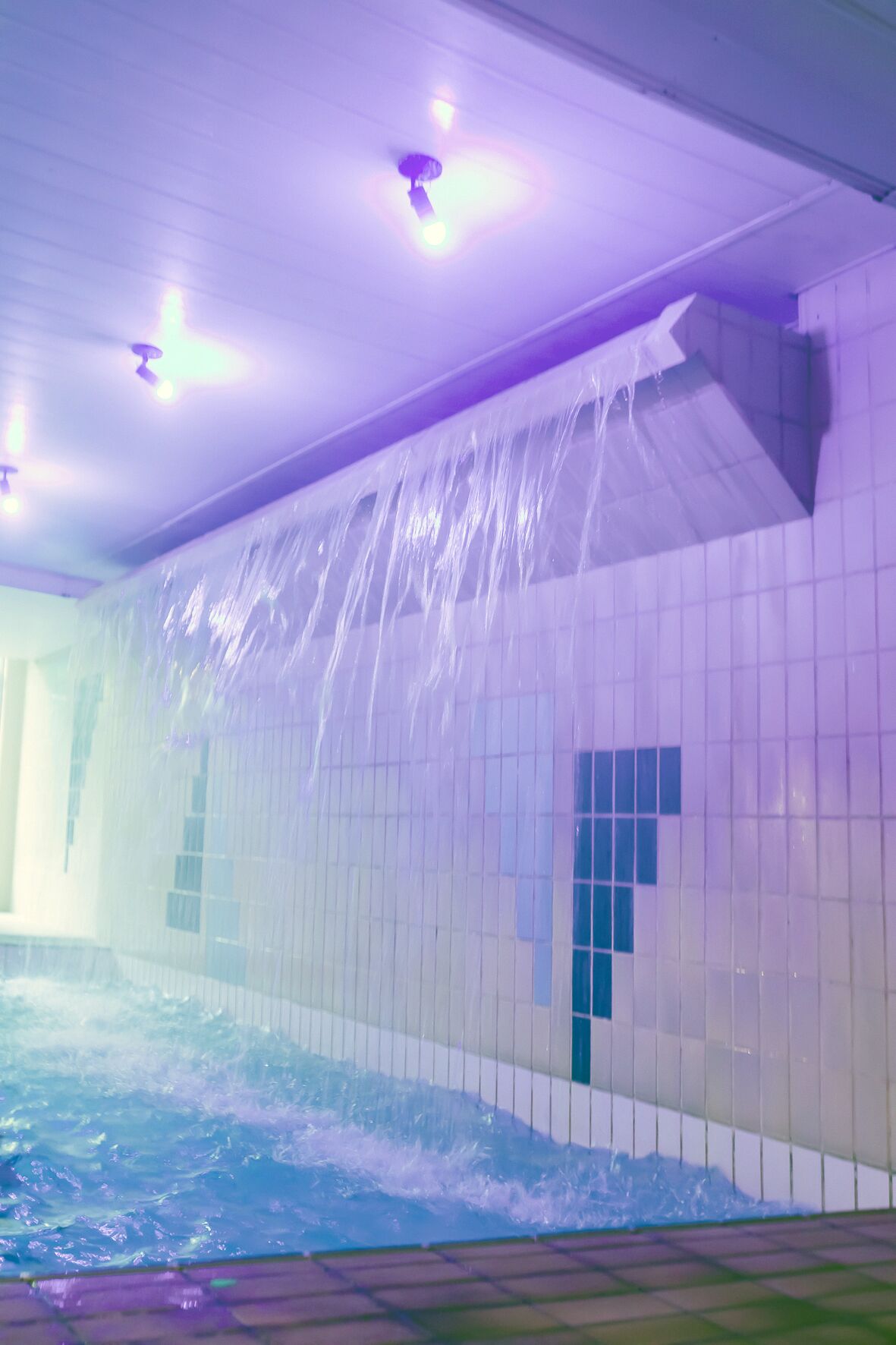Una piscina con caída de agua, paredes con azulejos e iluminación en el techo.