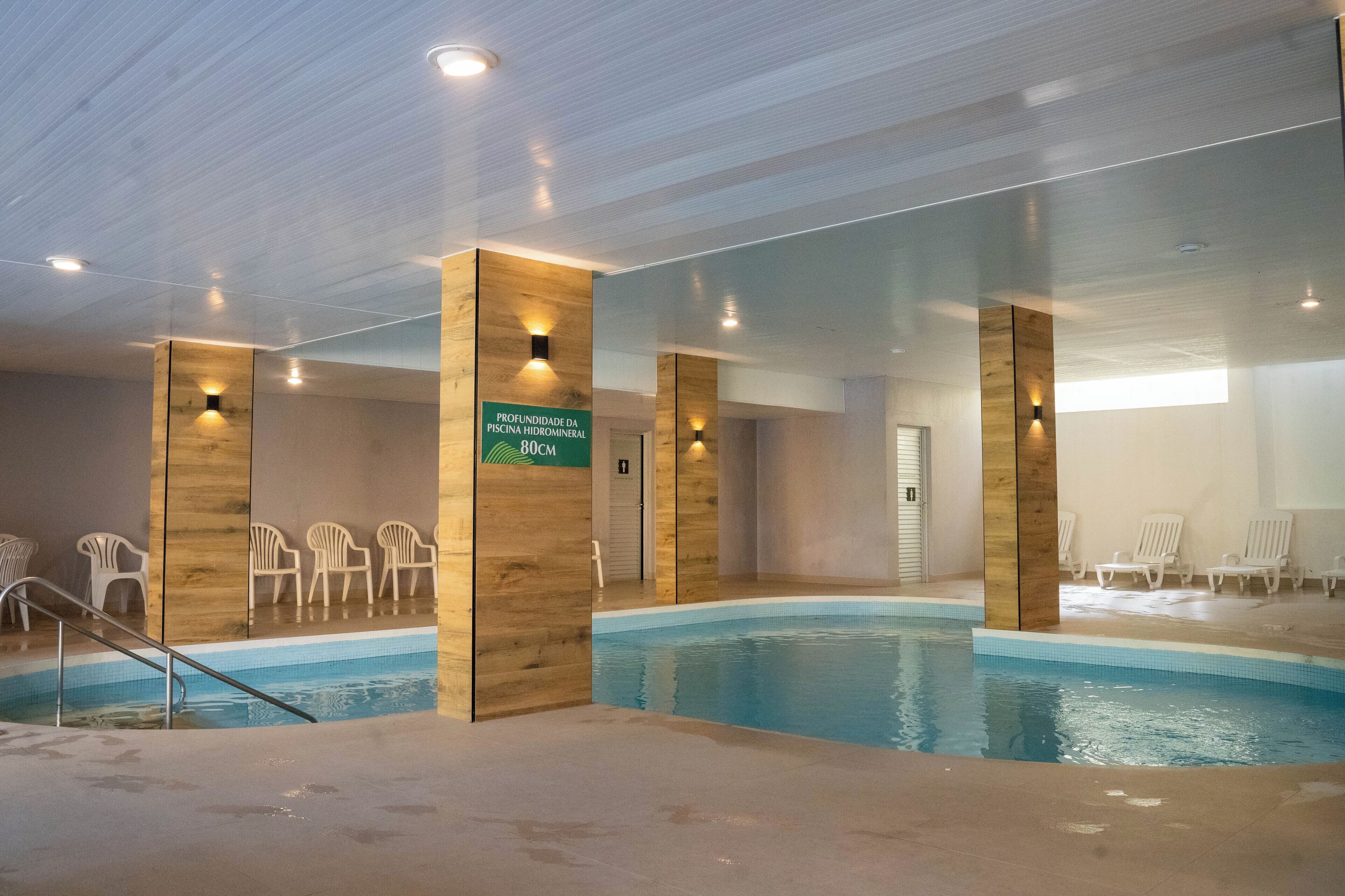 Piscina interna com pilares de madeira, cadeiras brancas e uma placa indicando a profundidade da piscina.