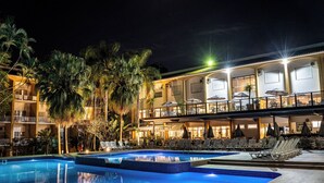 A nighttime swimming pool area with lounge chairs and umbrellas, surrounded by palm trees and a building with balconies.