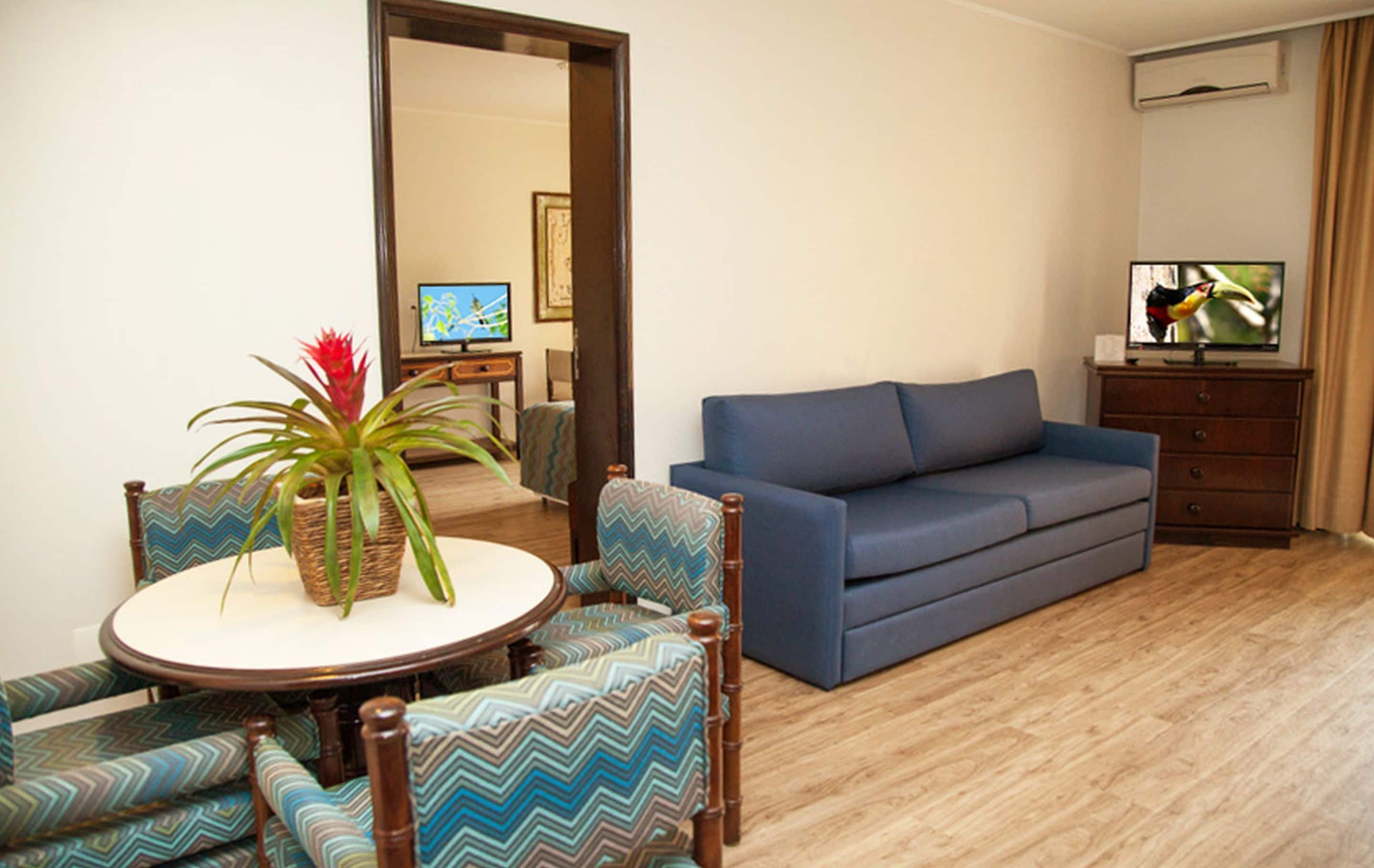 A living room with a blue sofa, a round table with chairs, a potted plant, and a television.