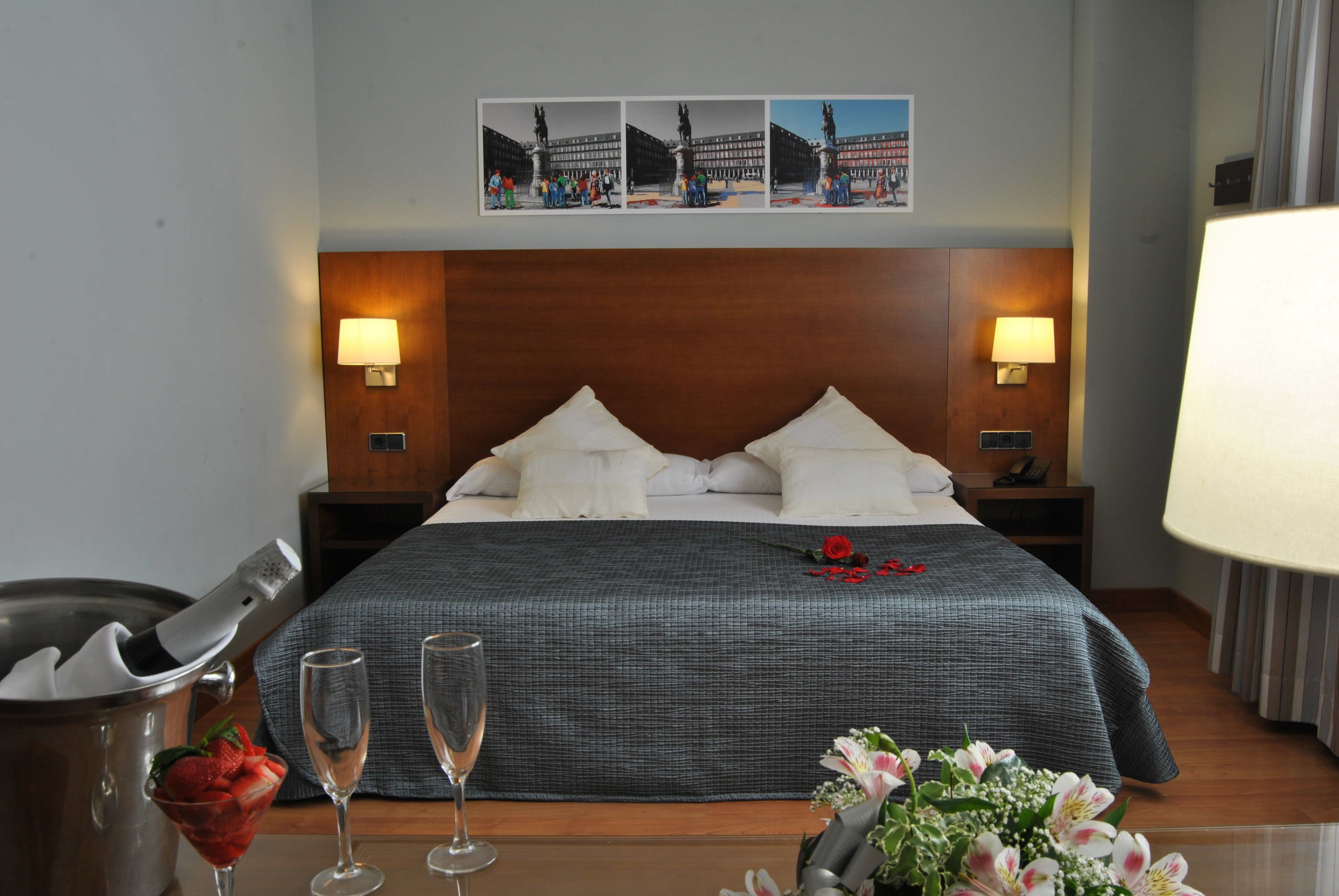 Een hotelkamer met een groot bed, twee bedlampen, een nachtkastje, een bijzettafeltje met een glas water, een schaaltje aardbeien en een bloevas.