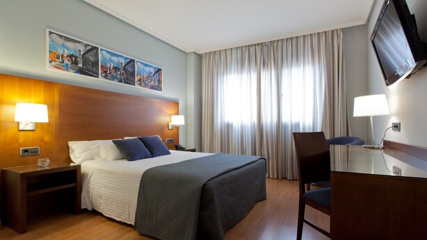 A hotel room with a bed, two bedside tables with lamps, a desk with a chair, a TV mounted on the wall, and a large window with curtains. - Hotel Porcel Avant (Torrejón de Ardoz)