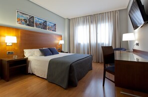 Quarto de hotel com uma cama, duas mesas de cabeceira com luminárias, uma escrivaninha com cadeira, uma TV fixada na parede e uma grande janela com cortinas.